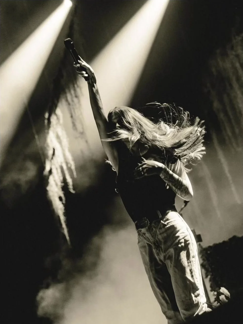 In black and white: Ethel Cain arched back with the microphone held straight up, her hair thrown across her face, two hard shafts of light crossing the ivy behind her. 2026. Frame from footage by @unicornsprinklessglitter, © the uploader.