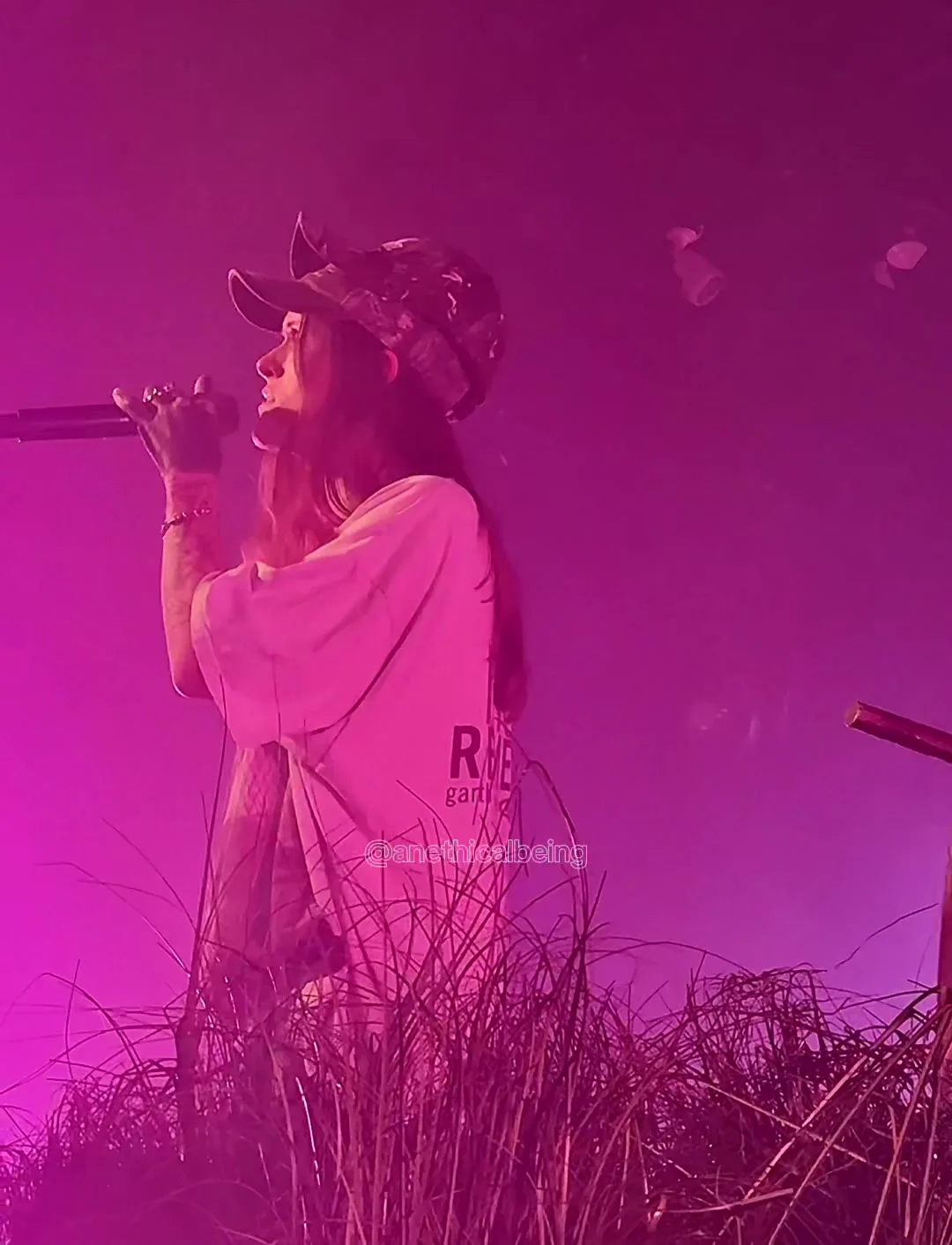 Ethel Cain backlit magenta in a cap and a loose white shirt, the microphone held high, the room hazed to a blur. Tallahassee · the second night. Frame from footage by @anethicalbeing, © the uploader.