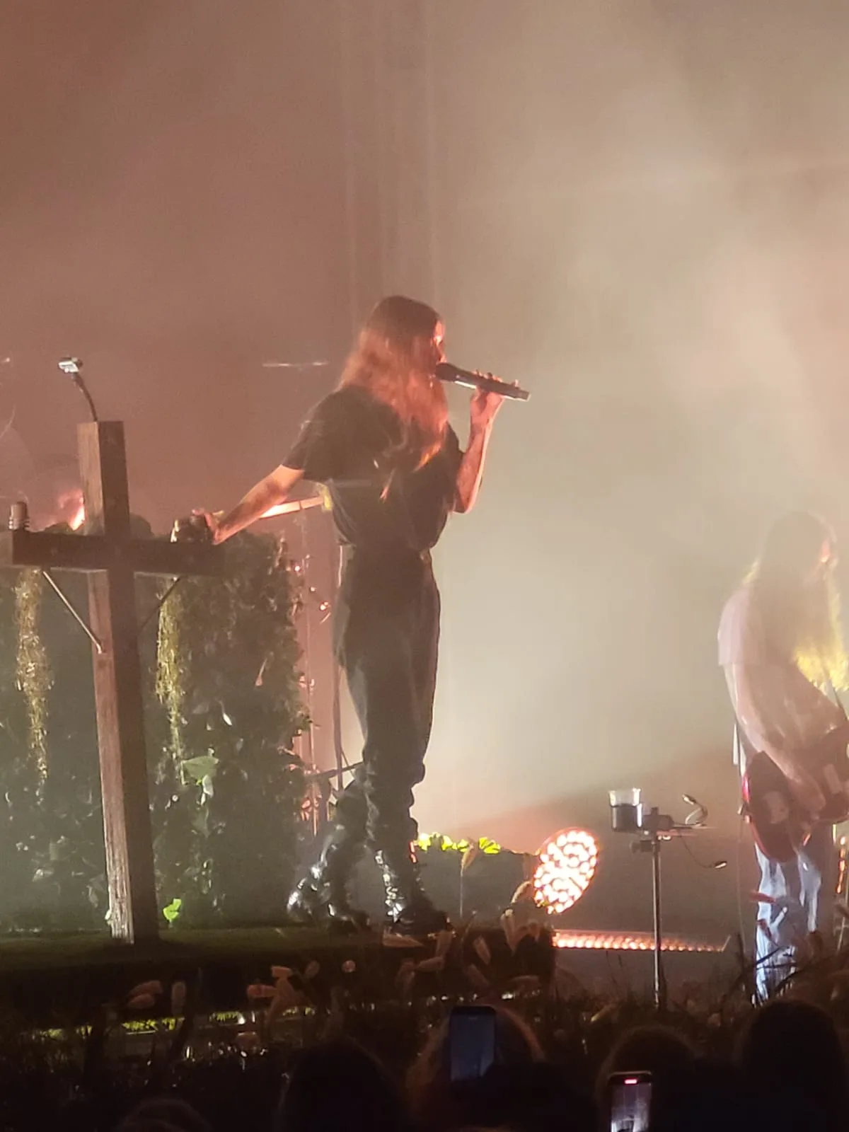 Ethel Cain turned away from the crowd with a hand on a mossed wooden cross, the stage full of amber smoke, a guitarist to her right. Lucerna, Prague · 1 November 2025. Photograph by TT2106, CC BY-SA 4.0.