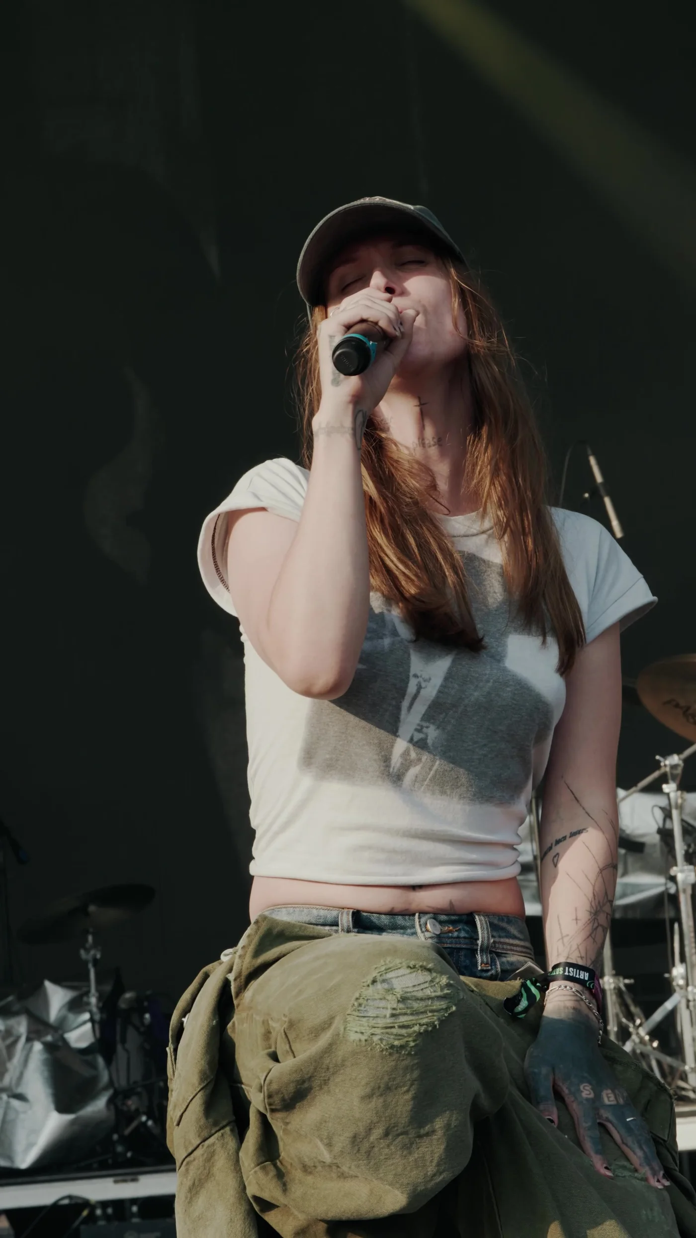 Ethel Cain in a cap and a white tee, eyes shut, singing into a handheld microphone against a black stage. Lollapalooza, Chicago · August 2024. Frame from footage by @lollapalooza, © the uploader.