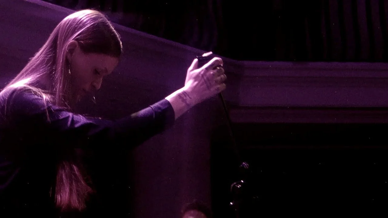 Ethel Cain reaching up against a dark wooden wall, lit low and violet, near the end of the European nights. Silent Green, Berlin · 1 December 2022. Frame from footage by thevinylsaurus, © the uploader.