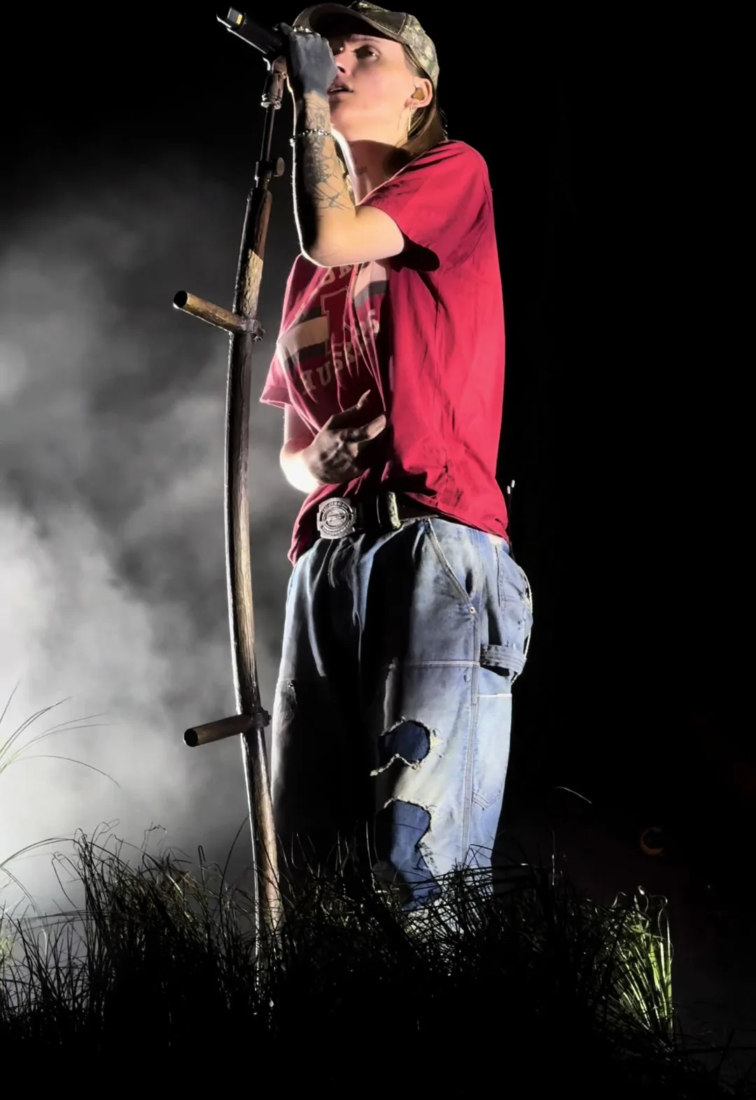 Ethel Cain in a red shirt and cap at the cross-shaped microphone stand, smoke rolling through the grass of the stage set. Cincinnati · 2025. Frame from footage by @casually_cassey, © the uploader.