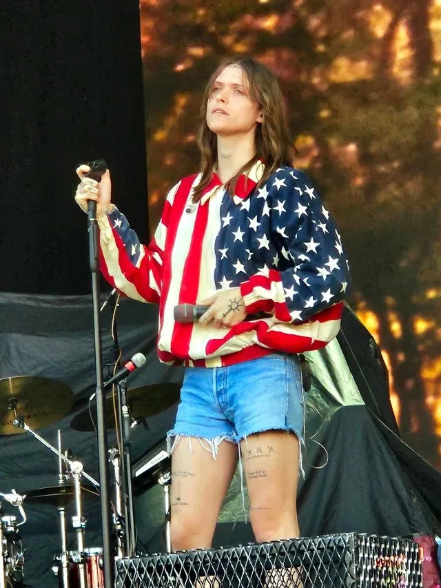 Ethel Cain at the microphone stand in a stars-and-stripes shirt and cut-off denim shorts, a wooded backdrop burning autumn-orange on the screen behind. Gunnersbury Park, London · 20 August 2023. Photograph by u/ishouldgooutmore on Reddit, CC BY 4.0.