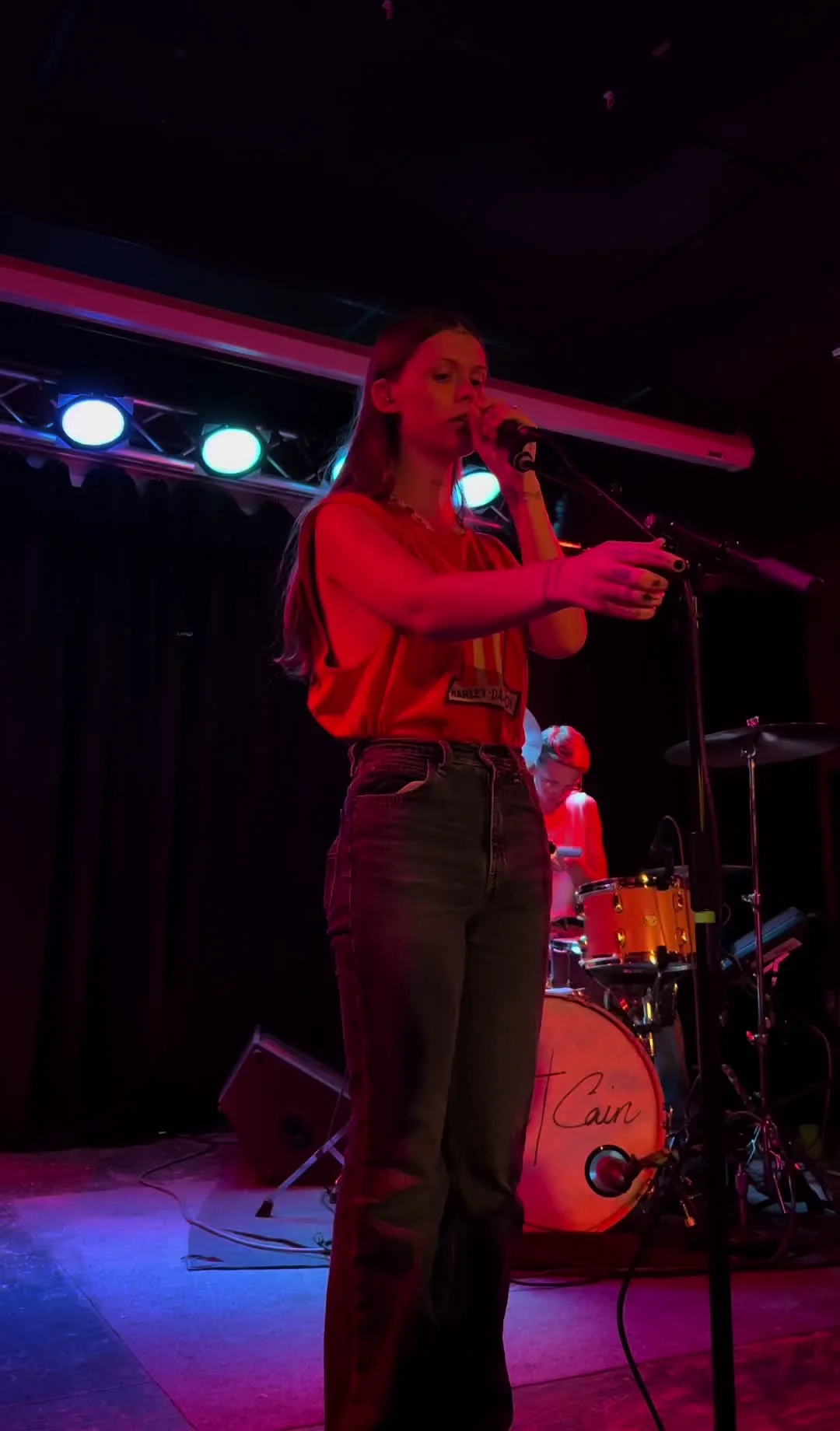 Ethel Cain in a red shirt on a low stage in a small room, her own name on the drum head behind her. the opening night · July 2022. Frame from footage by @_nicholashill, © the uploader.